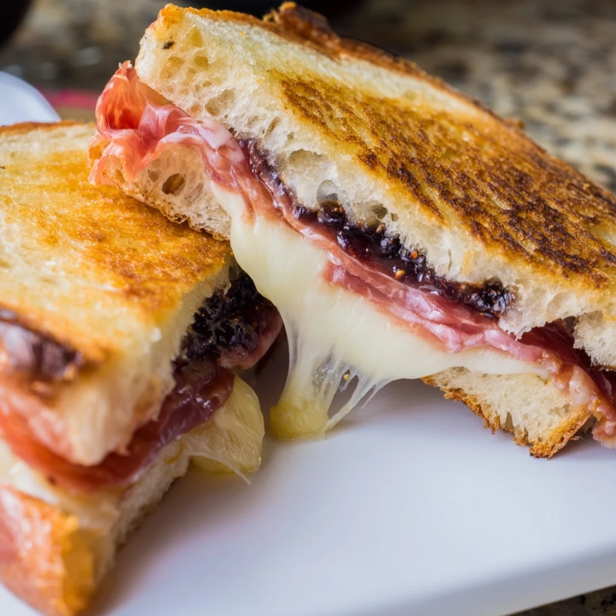 A close-up of a Crispy Prosciutto & Fig Panini with bubbly, golden-brown bread and sweet fig jam oozing from the edges.  