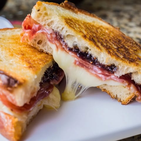 A close-up of a Crispy Prosciutto & Fig Panini with bubbly, golden-brown bread and sweet fig jam oozing from the edges.  